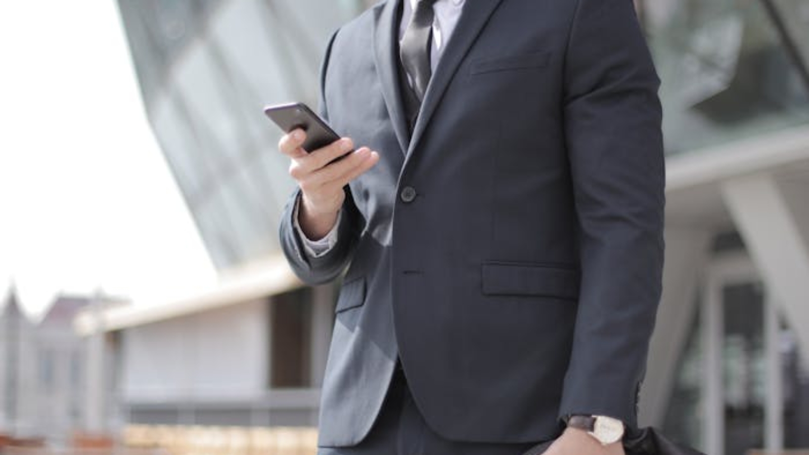 A confident businessman in a suit smiles while texting outside a modern building.