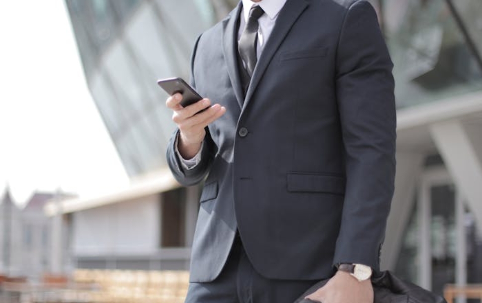 A confident businessman in a suit smiles while texting outside a modern building.